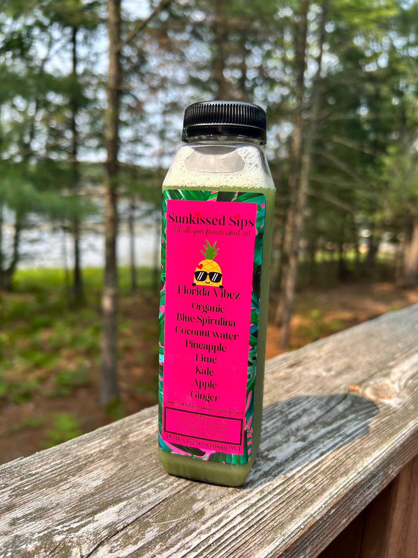 A clear plastic bottle of juice with a black cap stands on a wooden deck railing outdoors. The bottle contains a pale green liquid and has a bright pink label reading 'Sunkissed Sips' and 'Florida Blue'.