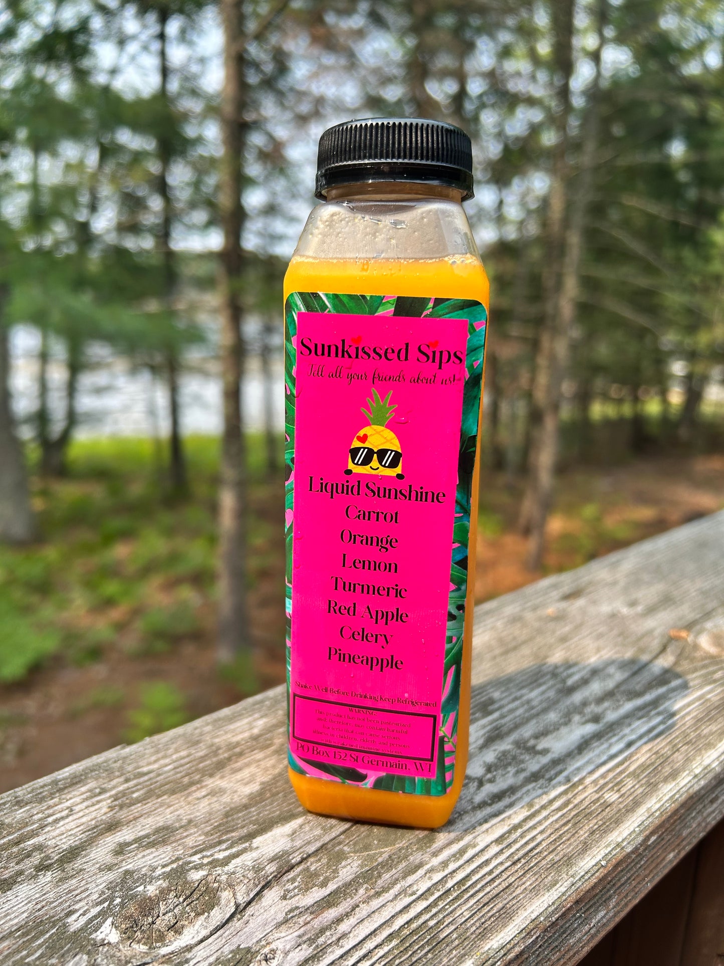A bottle of Sunkissed Sips Liquid Sunshine juice with a black cap and a bright pink tropical label, standing on a weathered wooden railing with a blurred forest background.