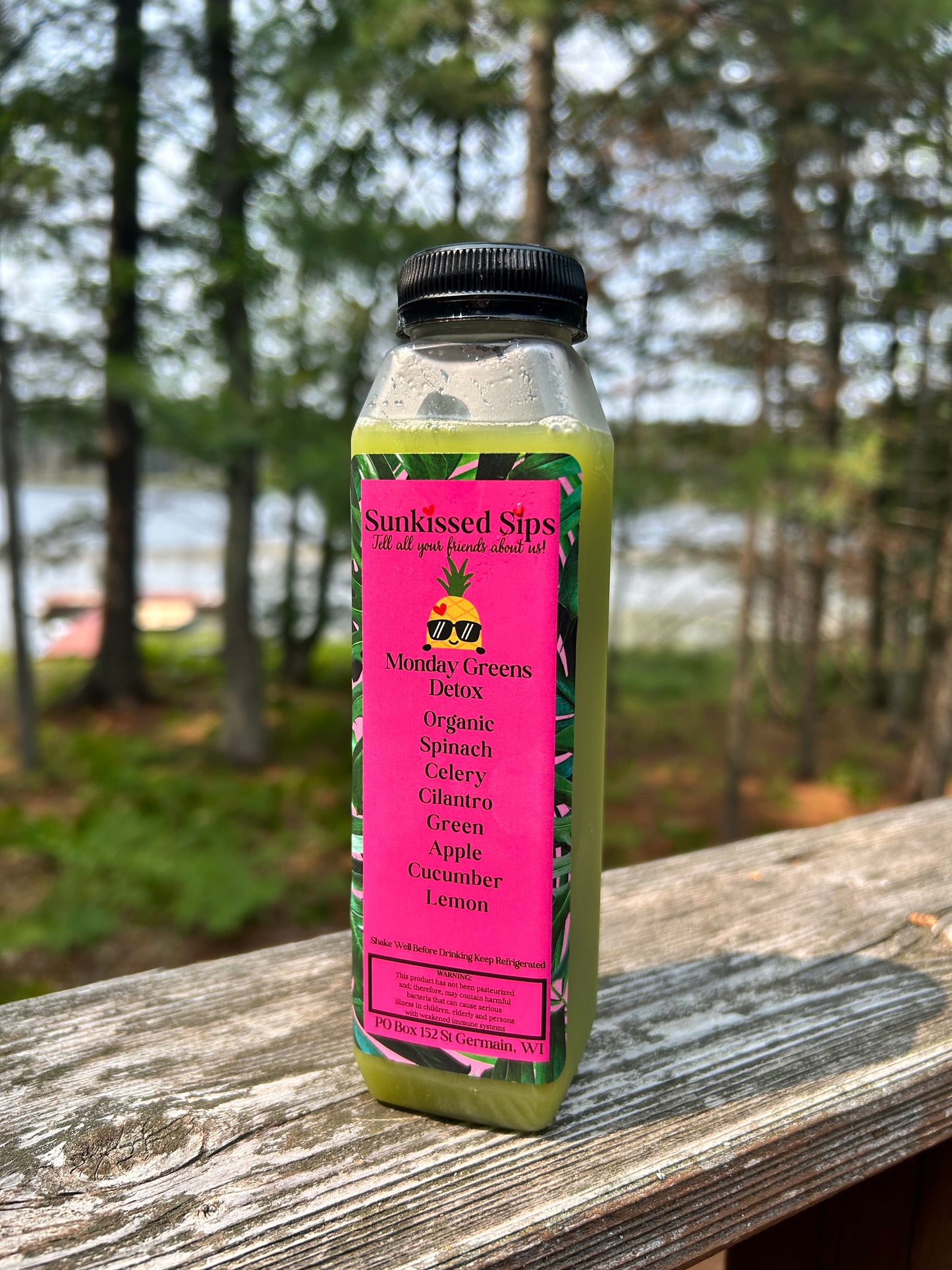 A bottle of Sunkissed Sips 'Monday Greens' detox juice with a bright pink label, sitting on a wooden deck railing with a blurred forest background.