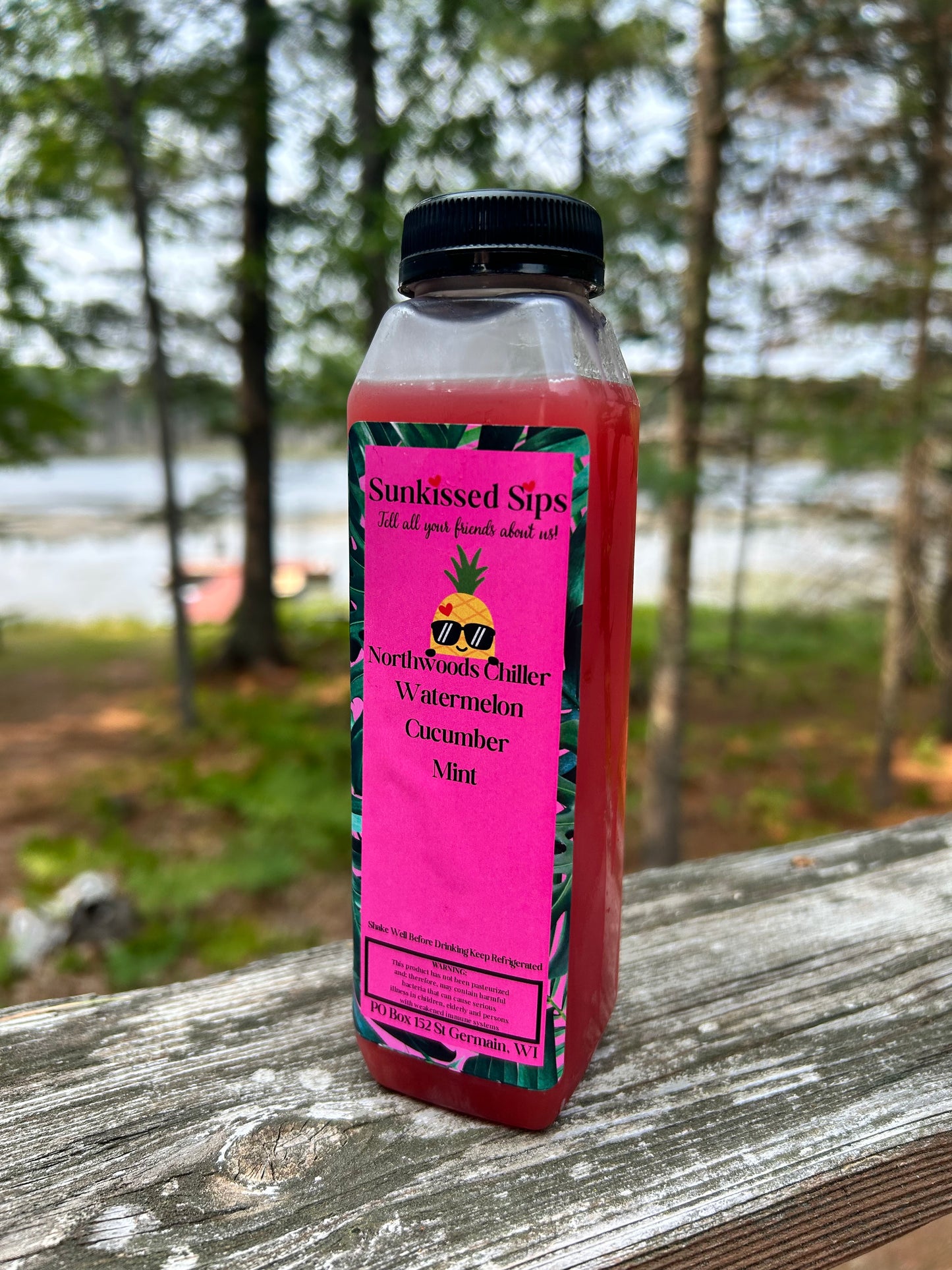 A bottle of Sunkissed Sips Watermelon Cucumber Mint juice sits on a weathered wooden railing with a blurred outdoor forest and lake background.