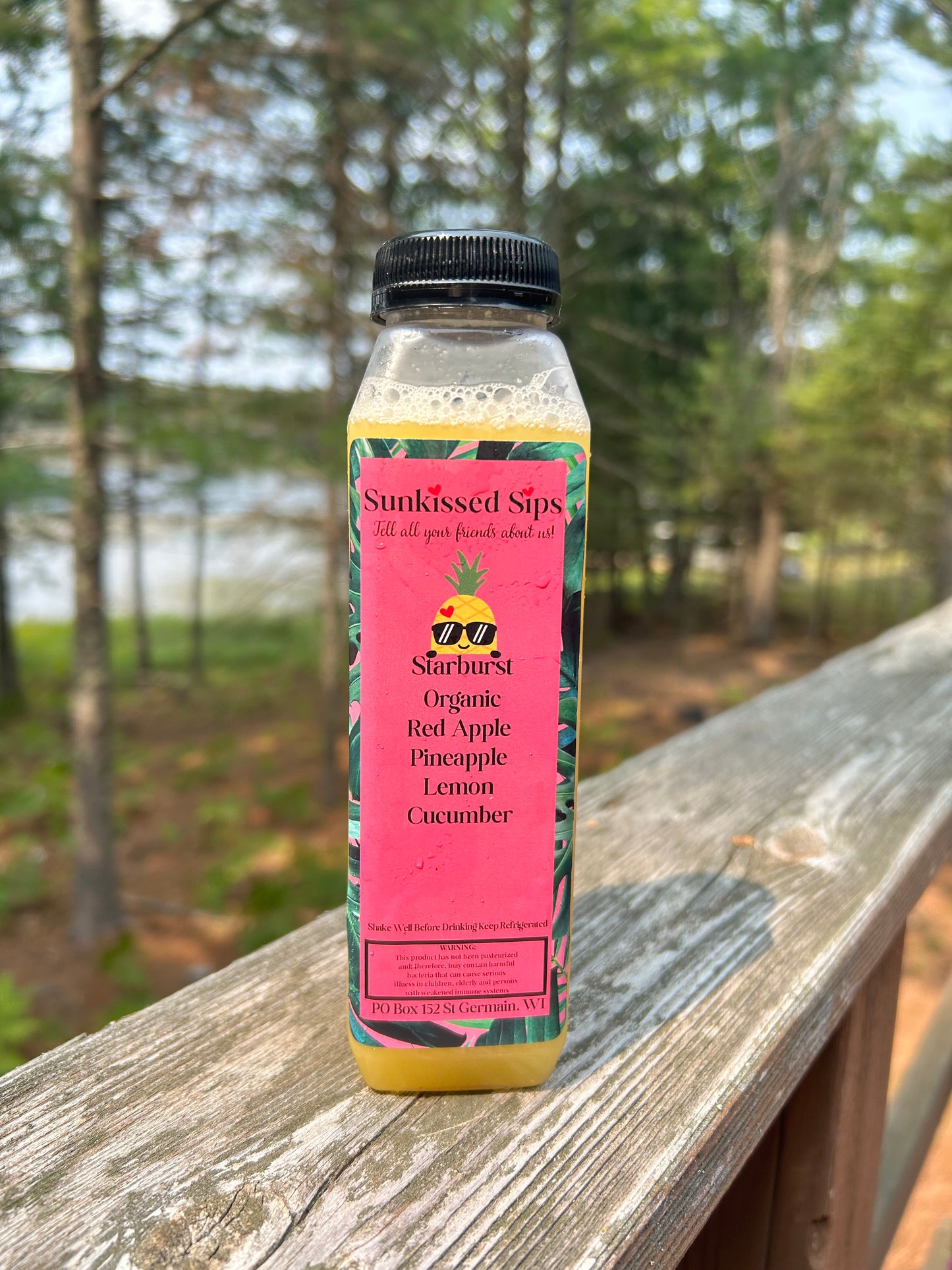 A bottle of Sunkissed Sips Organic Red Apple, Pineapple, Lemon, and Cucumber juice sits on a wooden deck railing with a blurred forest background.