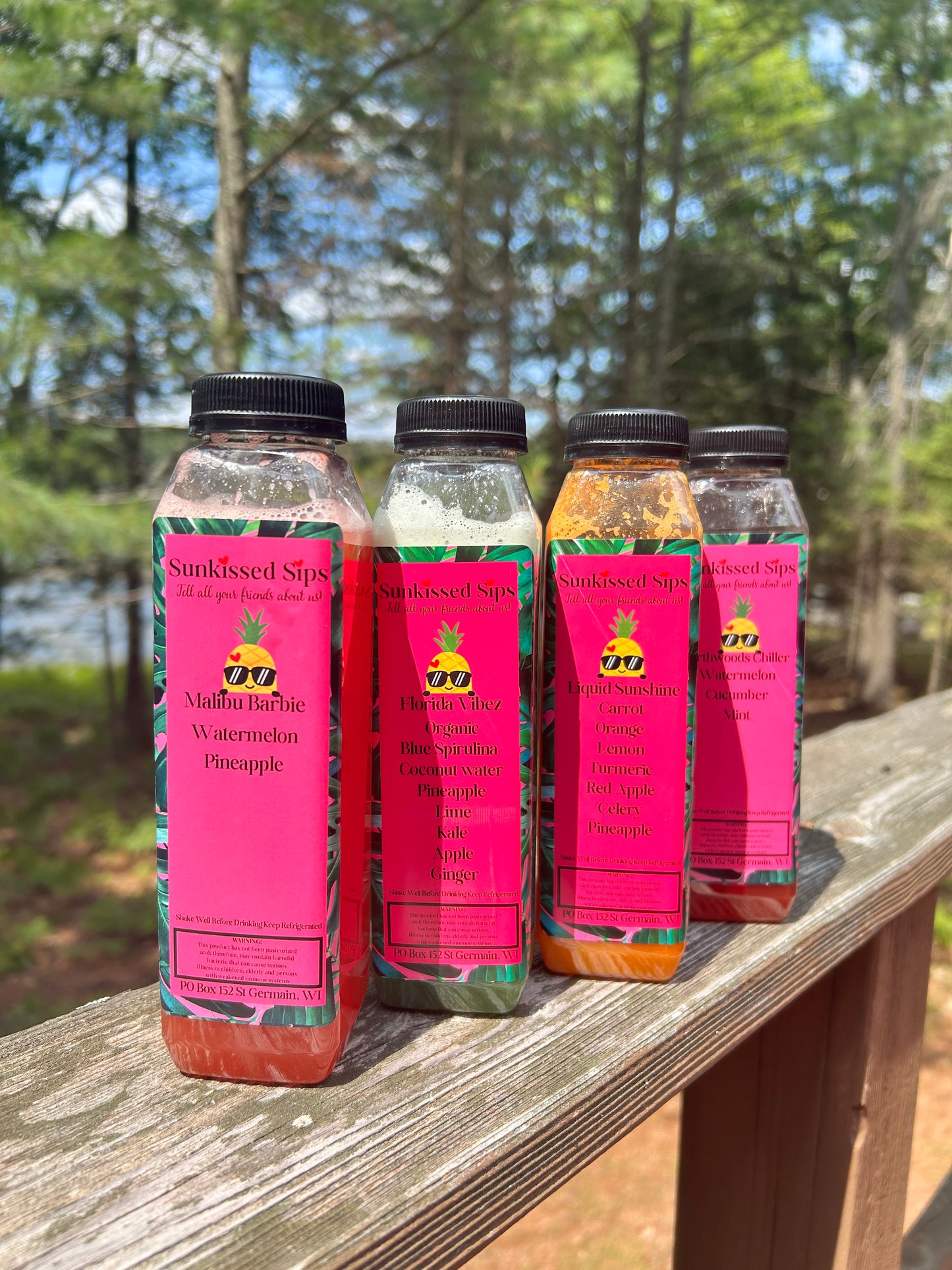 Four bottled juices from SunKissed Sips are lined up on a wooden deck railing outdoors with a blurred forest background. The bottles contain pink, green, orange, and red juices and feature bright pink labels with tropical designs.